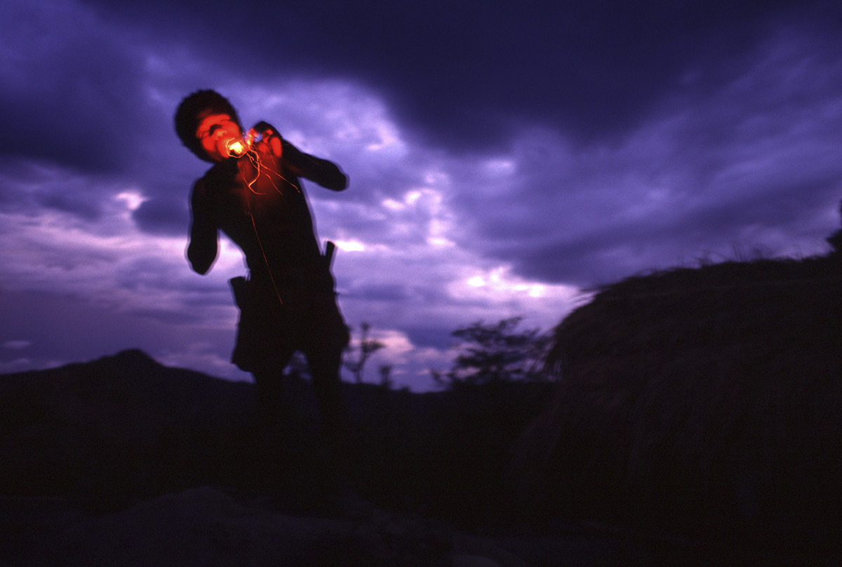An Aetas man blows on grassy fire in dusk scene on the dusty road to the remote village of Pili in Zambales. Philippines, 1999