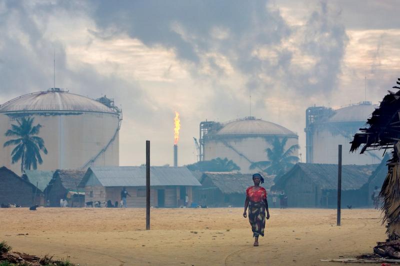 Photo: A woman passes the dirt soccer field and town square in the fishing village of Finima, which is a newly relocated community caused by the rapid growth of the Nigerian Liquified Natural Gas plant on Bonnie Island. Niger Delta, Nigeria 2006 Archival Pigment Print #3041