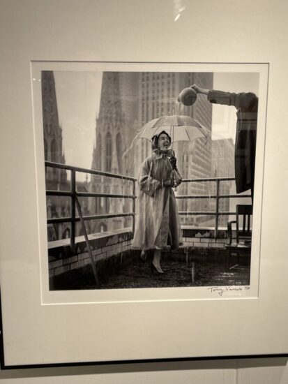 snapshot of a framed photograph showing a fashion model on a balcony with water being poured over her umbrella with NYC skyline in background
